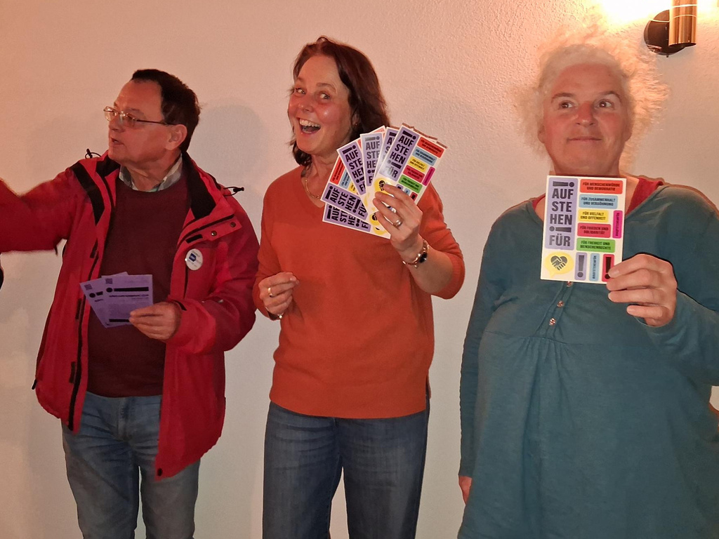 1 Mann und 2 Frauen, die Sticker-Postkarten der Aktion "Aufstehen für ..." zeigen